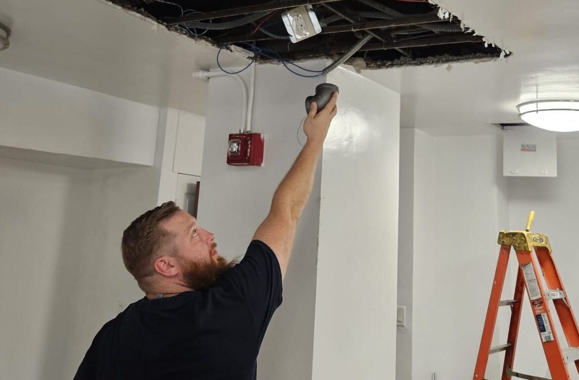 Ariel Services, Inc. Plumber holding pipe elbow, repairing gas piping in ceiling