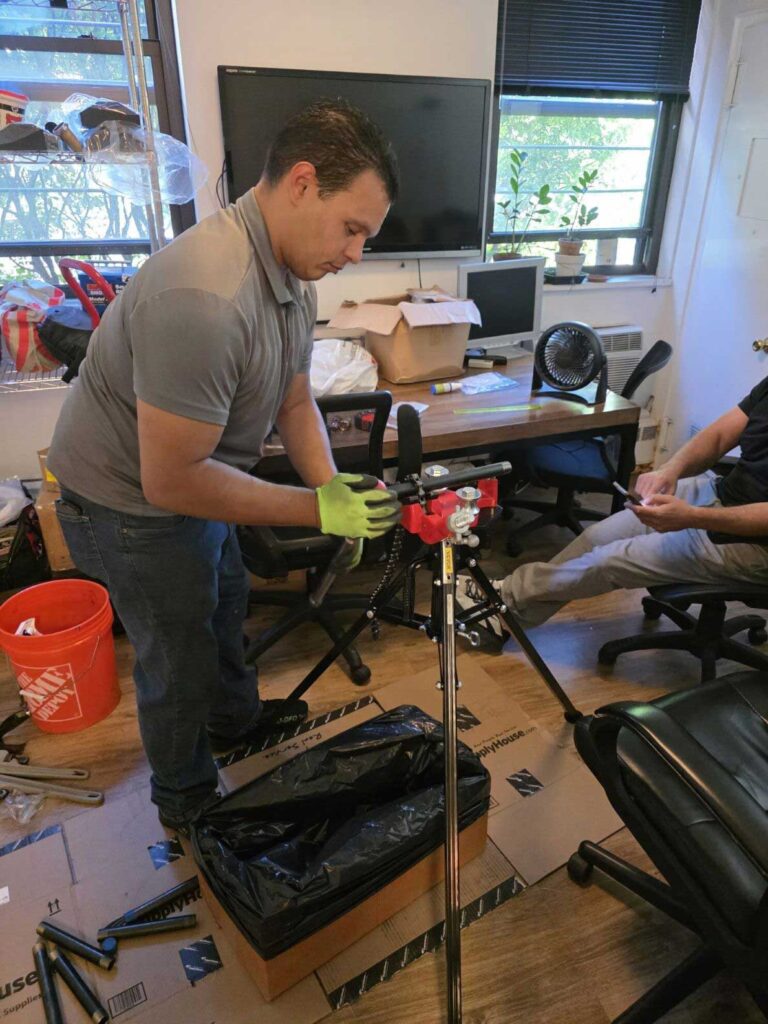 Technician assembling a pipe threading machine on cardboard in an office, Ariel Service Inc, with a coworker seated nearby.