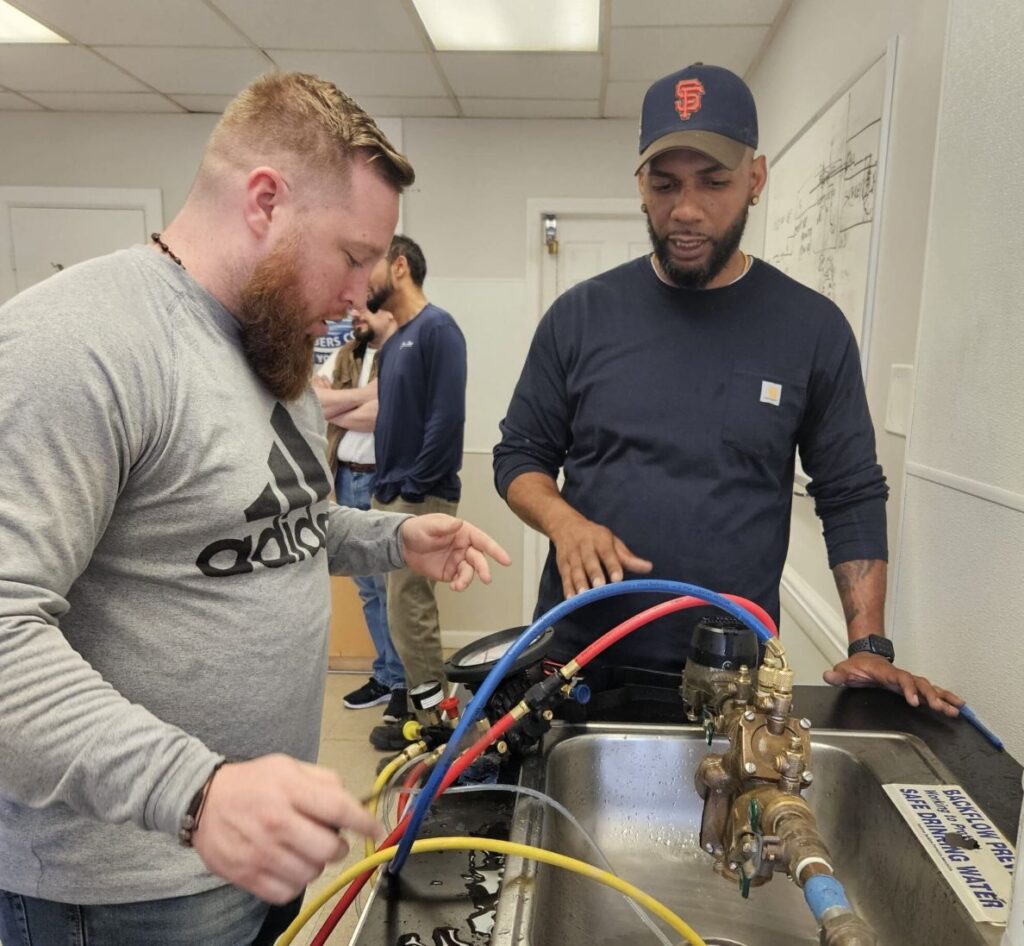 Two technicians testing a backflow preventer over a sink with gauge hoses attached, Ariel Service Inc, in a training classroom setting.