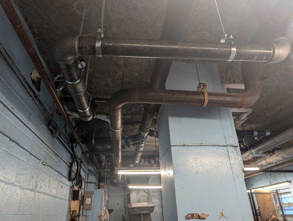 Basement ceiling with overhead steel and cast iron piping, Ariel Service Inc, running past a support column in a commercial building.