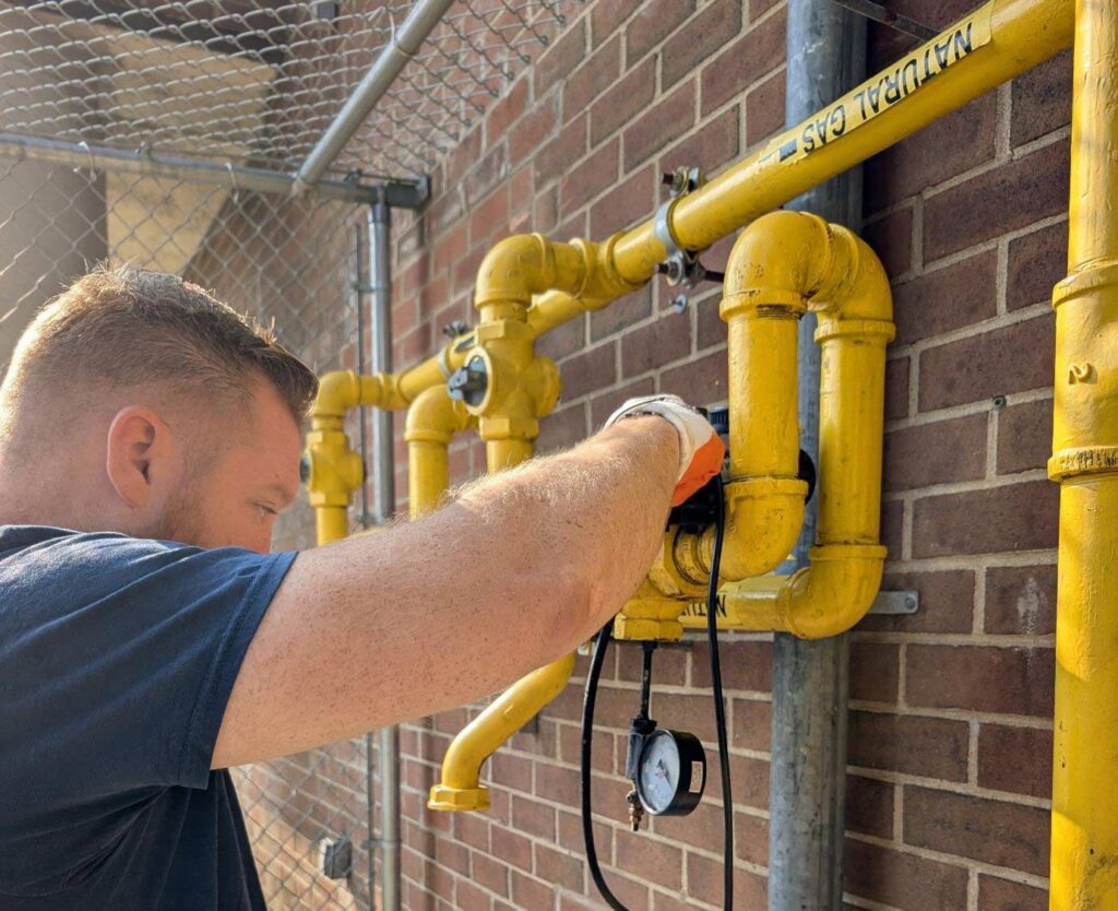 Technician adjusting a gas regulator on yellow-coated piping against a brick wall, Ariel Service Inc, with a pressure gauge attached below.