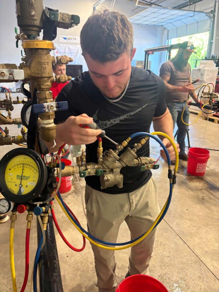 Technician using a screwdriver on a brass backflow preventer valve assembly, Ariel Service Inc, with manifold gauges and hoses attached during testing.